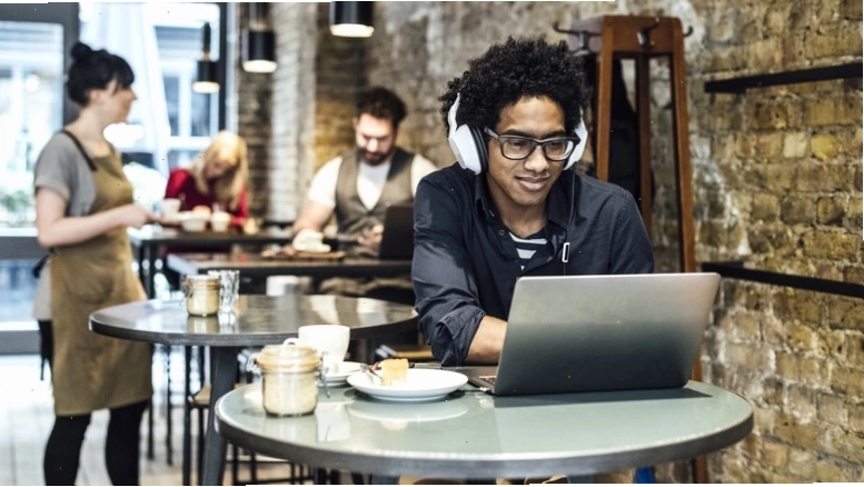 Người ngồi làm việc tại quán cafe với laptop và tai nghe, phía sau có khách hàng khác